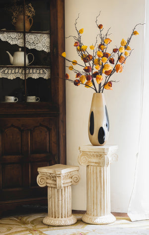 A tall, speckled vase with black spots, displayed on a classic column pedestal, holds branches with yellow and orange leaves in a bright interior.