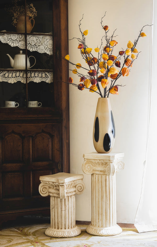 A tall, speckled vase with black spots, displayed on a classic column pedestal, holds branches with yellow and orange leaves in a bright interior.