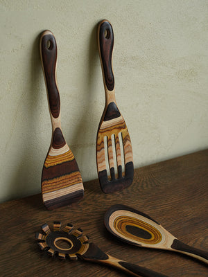 A collection of artisanal wooden kitchen tools with variegated brown grain designs, elegantly presented on a rustic wooden shelf against a plastered wall.