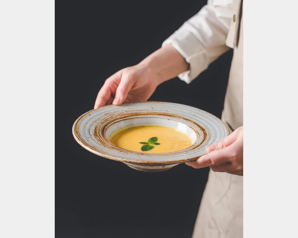 A person in a white shirt is serving a bowl of yellow soup with a mint leaf garnish. The Aphrodite's Casa logo appears in the top left corner.