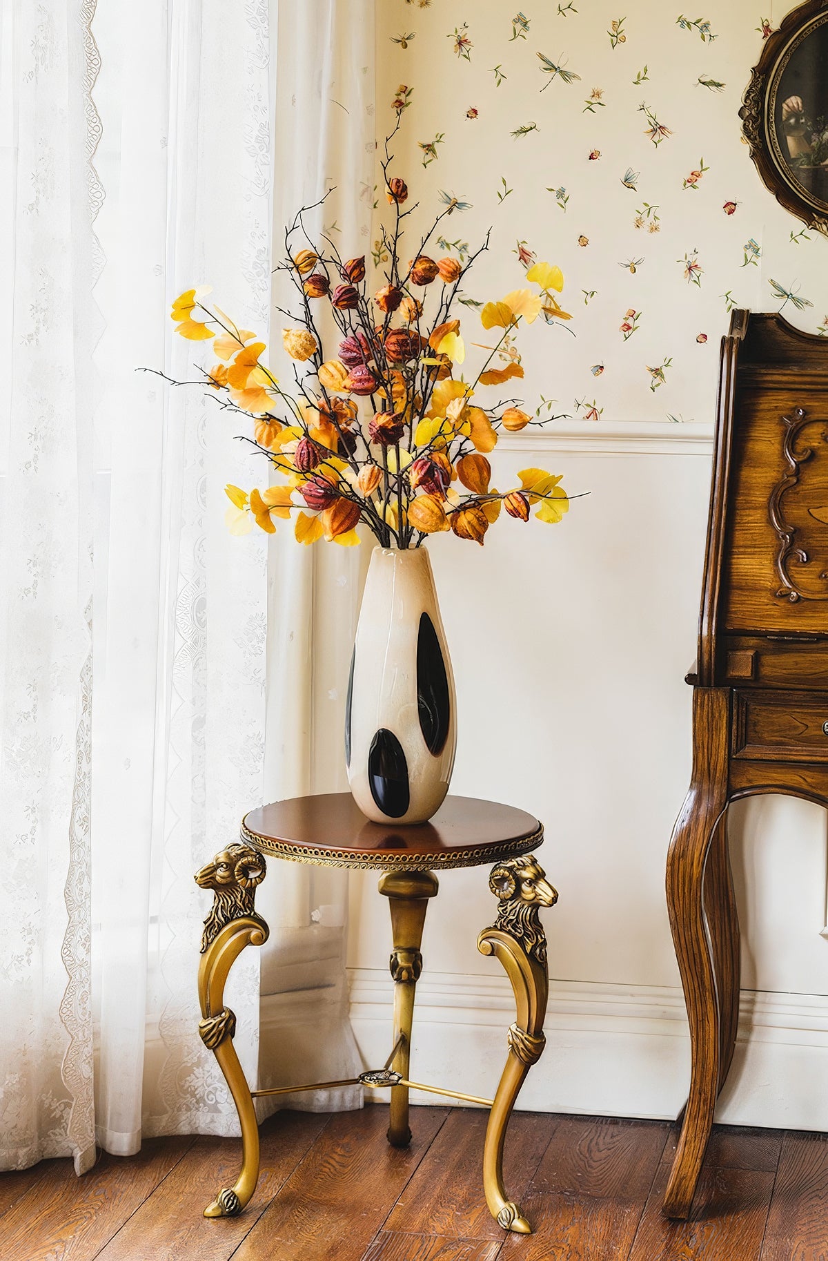 A uniquely shaped vase with black accents stands on an ornate golden-legged table, holding branches with vibrant autumn leaves, against a patterned wall.