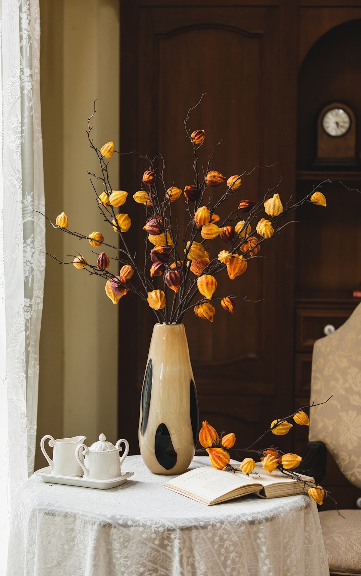 A vintage ceramic vase with black spots, placed on a wooden table, filled with branches bearing orange foliage, beside a classic teapot set.