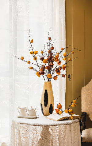 A vintage ceramic vase with black spots, placed on a wooden table, filled with branches bearing orange foliage, beside a classic teapot set