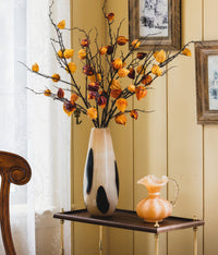 A speckled vase with bold black spots is situated on a round, gilded table, accompanied by an elegant, light peach-colored glass pitcher