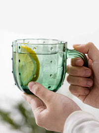 Green glass cup held in hands, with lemon water. | Grönt glas i händerna, med citronvatten