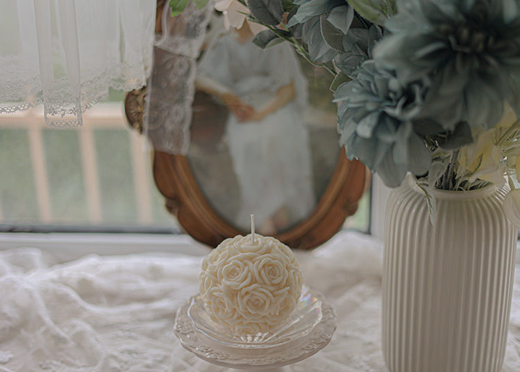 Elegant white rose candle, clear glass plate, floral arrangement backdrop. | Eleg. vit rosformad ljus, klar glastallrik, blomsterbakgrund.